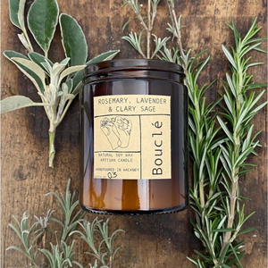 Soy wax aromatherapy candle in amber glass jar surrounded by sprigs of rosemary, lavender and clary sage representing it's essential oil scent blend.