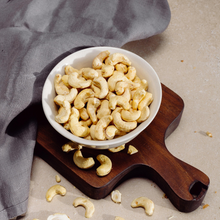 Load image into Gallery viewer, Cashew nuts in bowl on wooden tray in natural setting.
