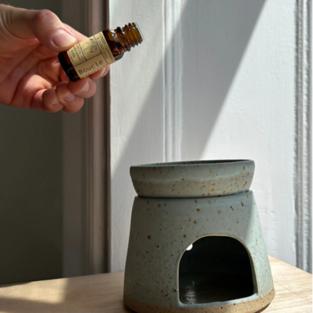 A hand holding a bottle of essential oil aromatherapy scent blend over an oil burner. Refill Mill.