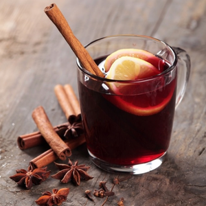 Glass mug of brewed mulled wine set on wooden surface with cinnamon logs and star anise adjacent and a cinnamon bark and lemon slices in the drink.
